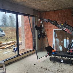 SKIBEN Skiba Przemysław - Montaż dużego okna z użyciem specjalistycznego robota na gąsienicach w surowym wnętrzu budynku. Widoczne przyssawki mocujące szybę.