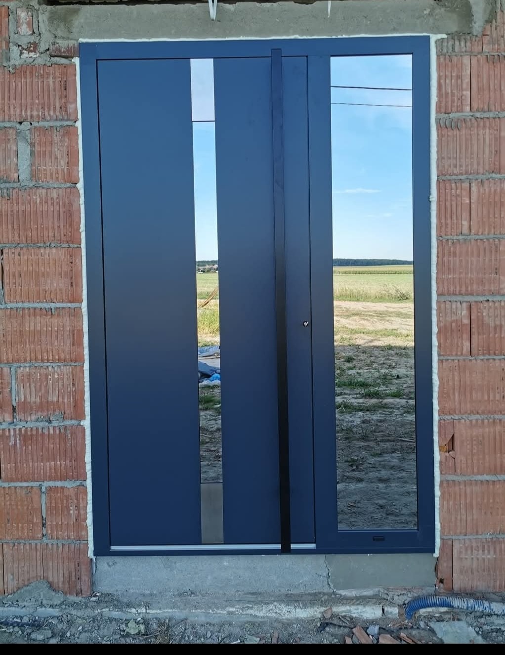 Zamontowane, grafitowe drzwi przesuwne z widokiem na pole, osadzone w murze z czerwonej cegły. Widoczne detale wykończenia i refleksy nieba w szybach.