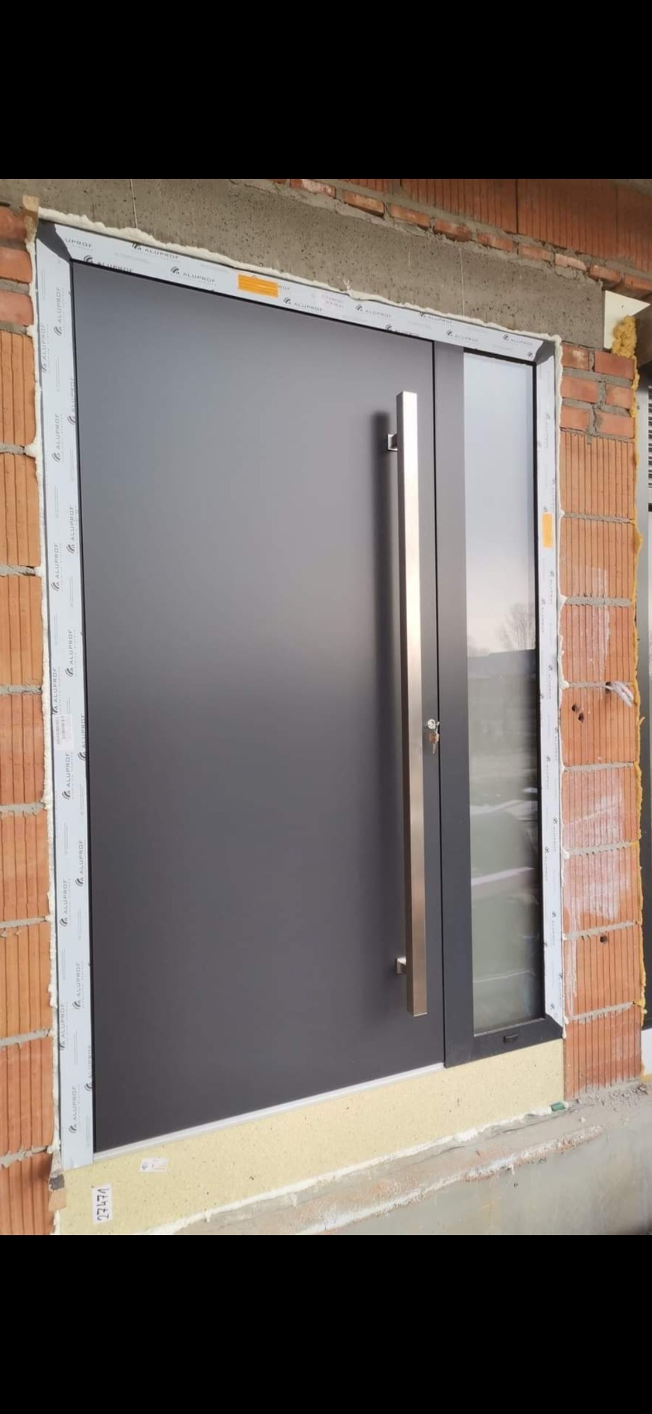 Nowoczesne, grafitowe drzwi zewnętrzne z długim, stalowym uchwytem i bocznym przeszkleniem, osadzone w surowym murze z widoczną izolacją.