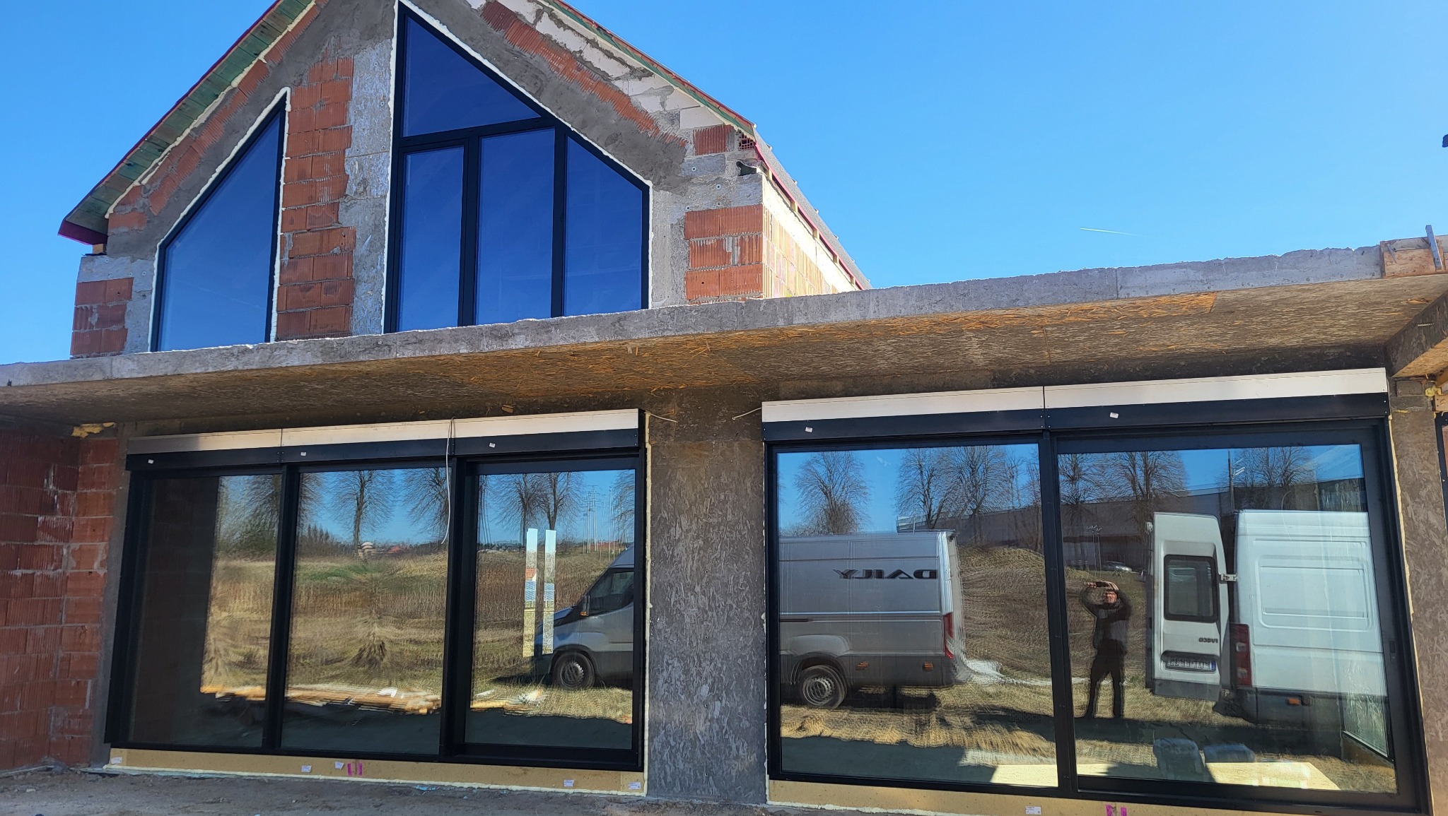 Surowe ściany domu w budowie z zamontowanymi oknami w czarnych ramach i roletami zewnętrznymi. W szybach odbijają się samochody dostawcze.
