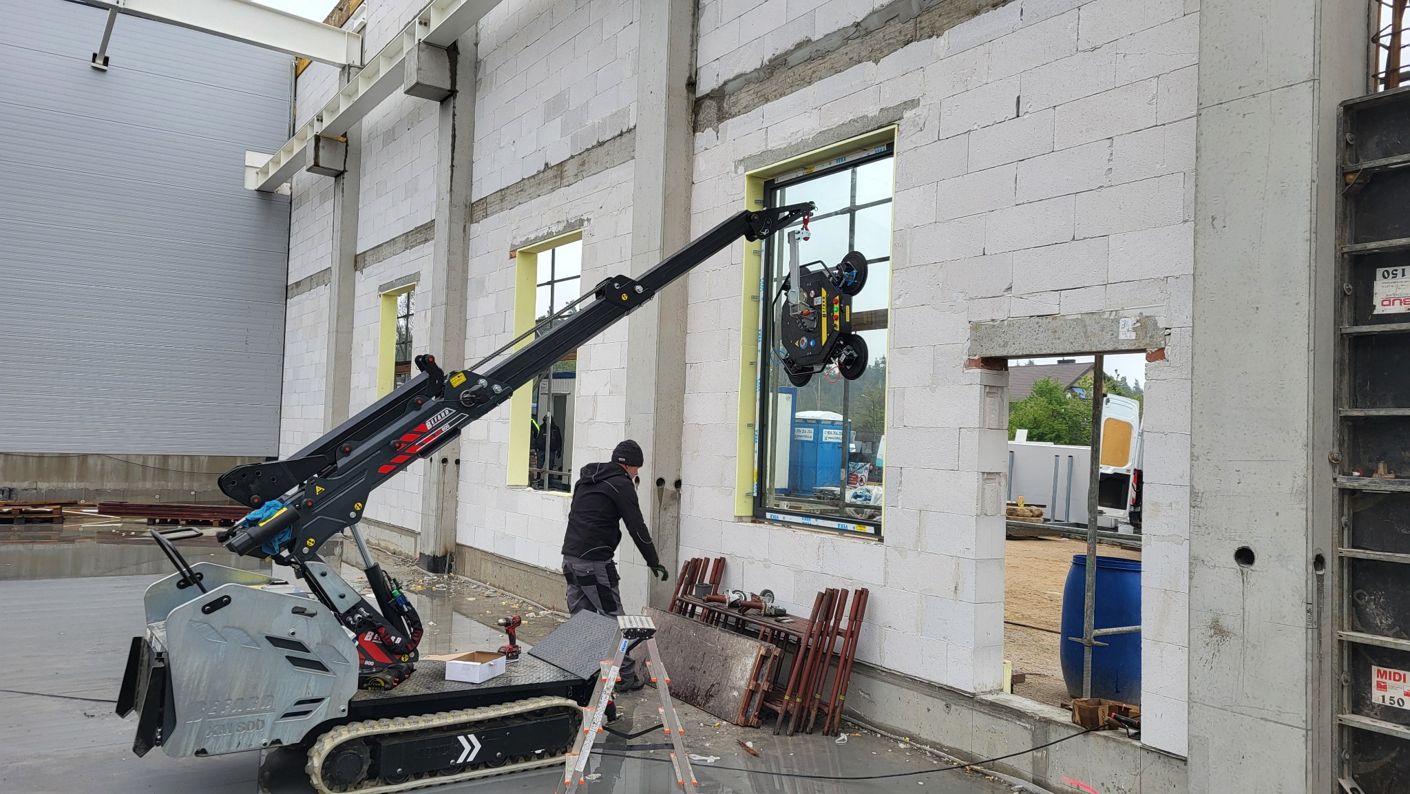 Montaż okna na budowie z użyciem specjalistycznego dźwigu przyssawkowego. W tle widoczne inne okna oraz elementy konstrukcyjne budynku.