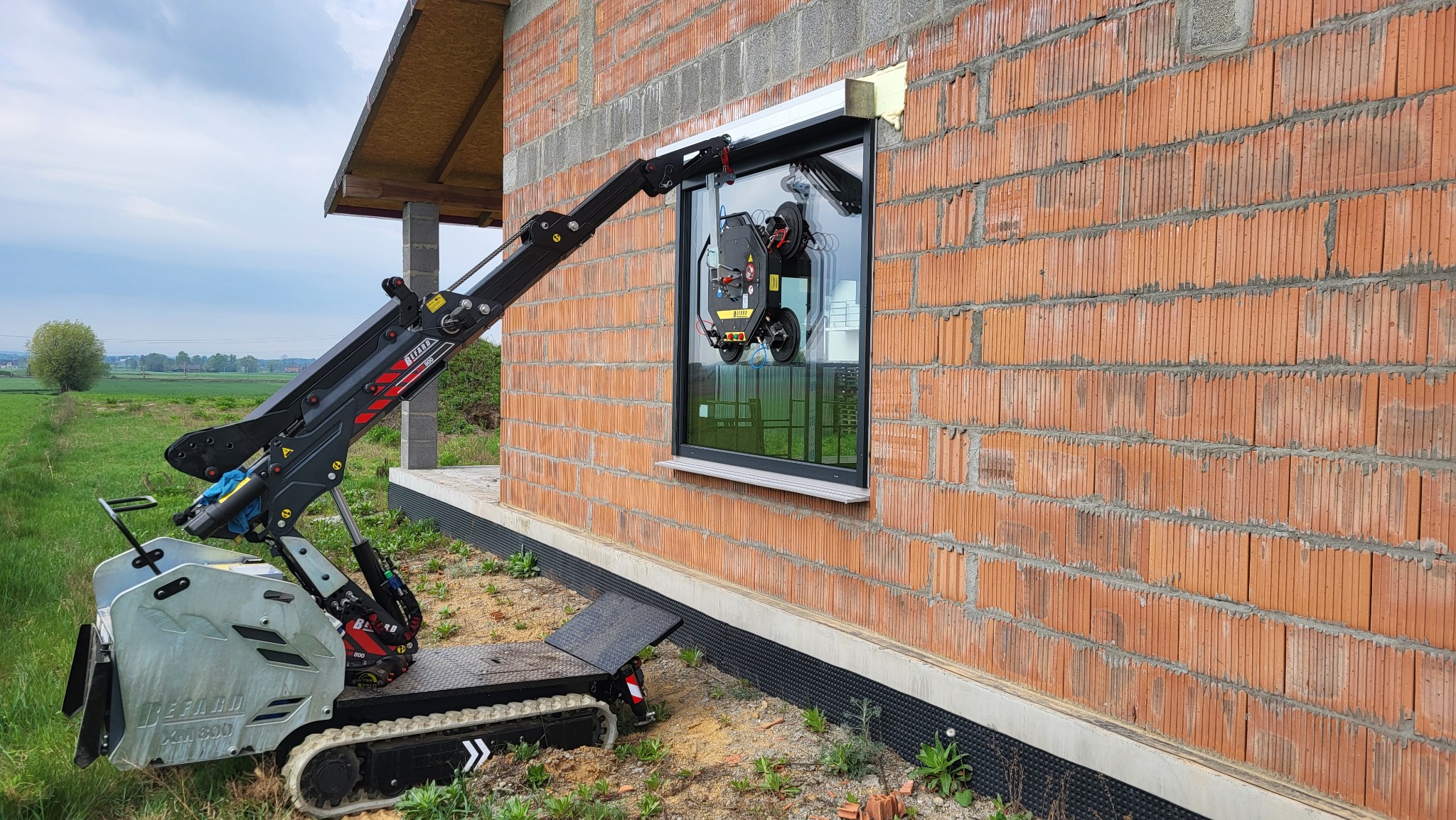 Czerwona ściana budynku w trakcie budowy z zamontowanym oknem. Do montażu użyto specjalistycznej maszyny z przyssawkami.