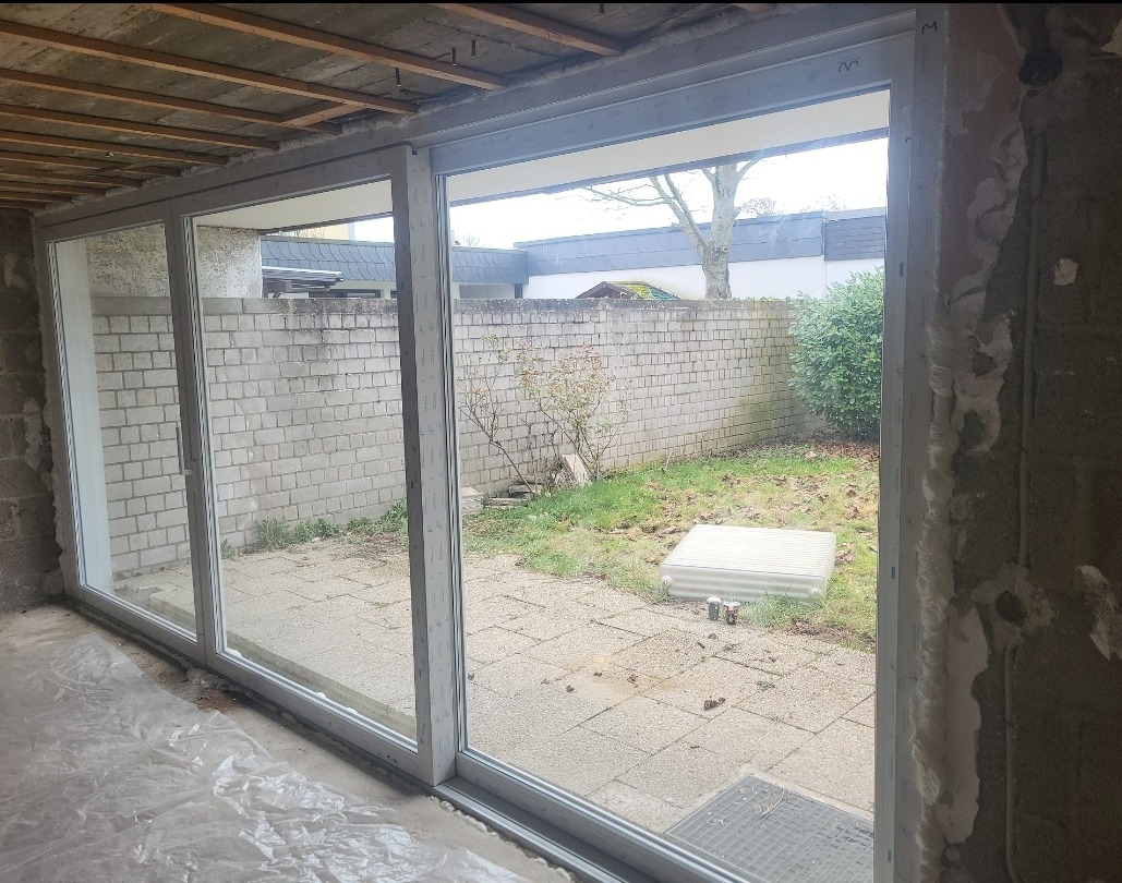 Montaż dużych, białych okien przesuwnych z widokiem na ogród. Surowy stan wykończenia wnętrza. Widoczne elementy konstrukcyjne sufitu i ściany.