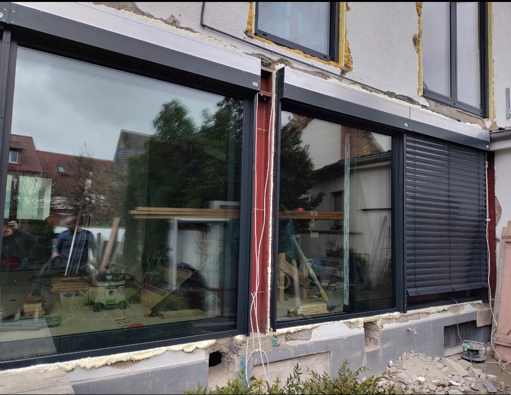 Montaż okien z roletami zewnętrznymi. Widoczne narzędzia pracy i ekipa montażowa. Okna w kolorze grafitowym, budynek w trakcie prac wykończeniowych.