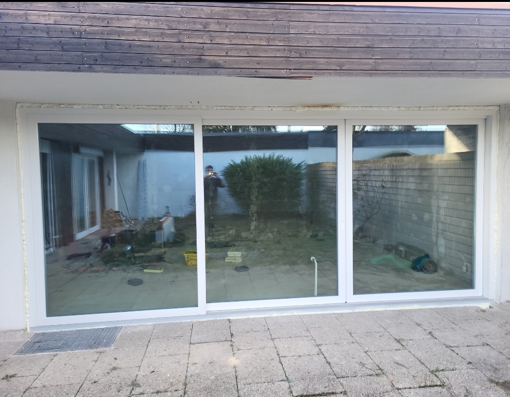 Zamontowane, trzyskrzydłowe drzwi przesuwne z białymi ramami, widok z zewnątrz na taras z kostki. W tle ogród.