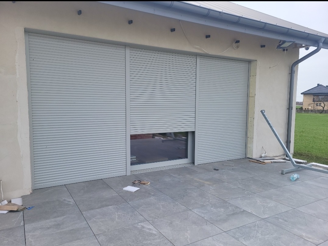 Zamknięte rolety zewnętrzne w kolorze szarym na oknie. Fragment otwartej rolety. Widoczne narzędzia montażowe i fragment budynku z szarą kostką.