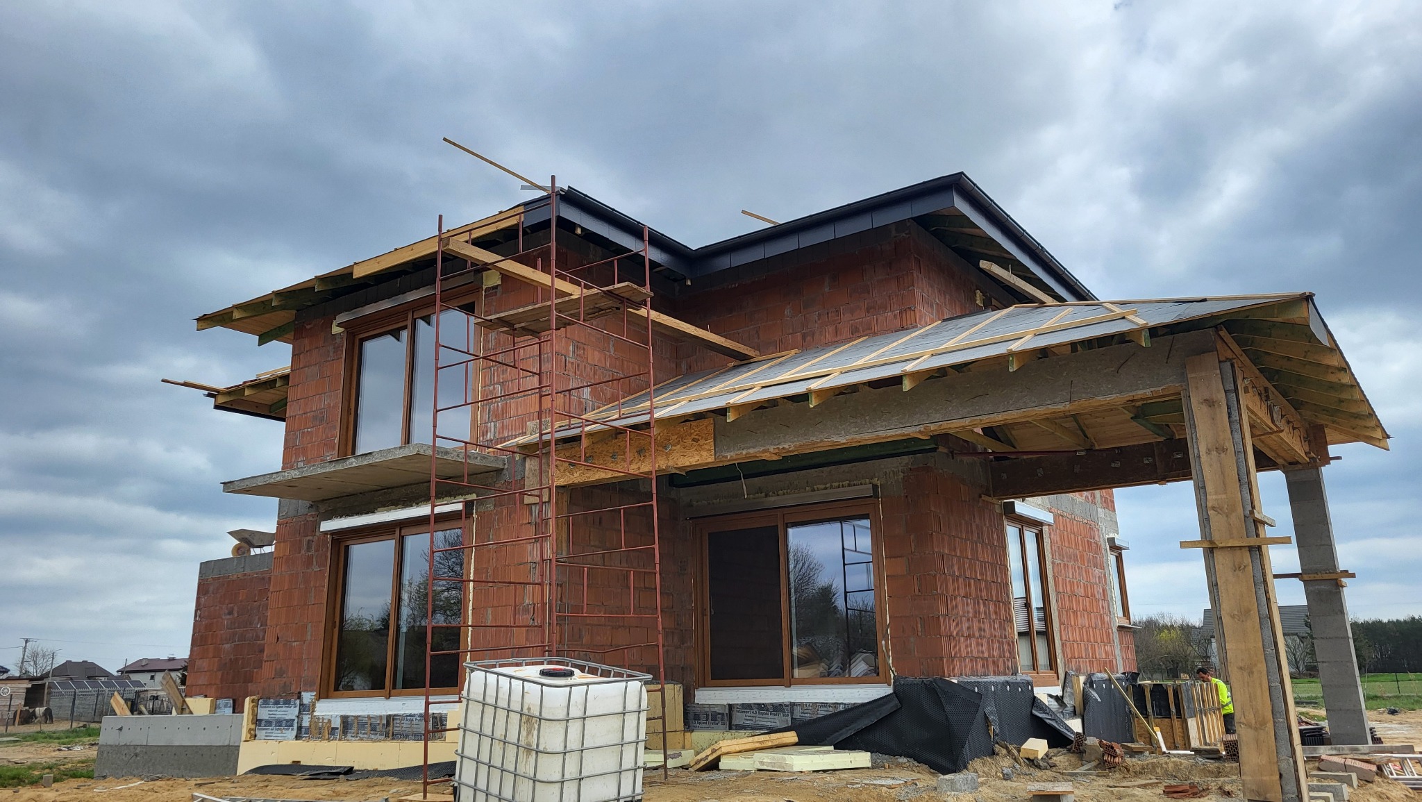 Budowa domu w trakcie: murowane ściany, okna z brązowymi ramami, rusztowania, drewniana konstrukcja dachu. Widok na budynek w surowym stanie.