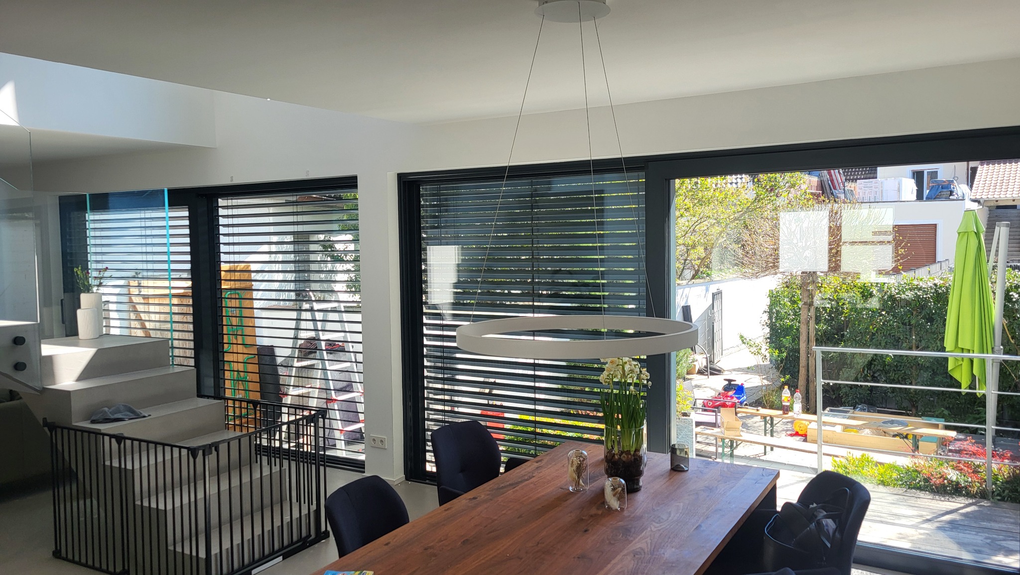 Nowoczesne, ciemne rolety zewnętrzne zamontowane w dużych oknach przesuwnych, widok z wnętrza domu na ogród w słoneczny dzień. Minimalistyczny design.