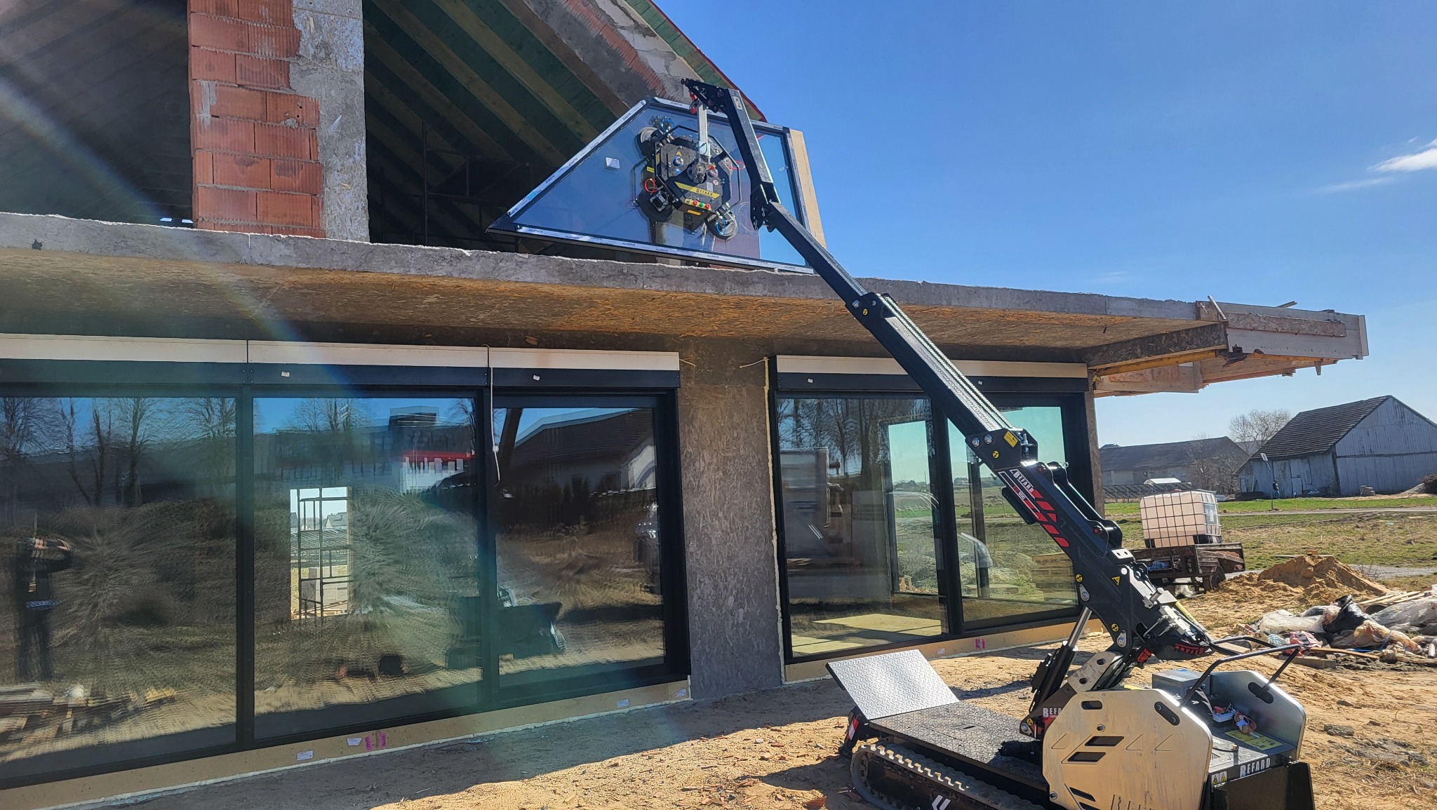 Montaż dużego okna za pomocą specjalistycznego dźwigu na placu budowy. Widoczne zainstalowane okna przesuwne i surowy stan budynku w tle.