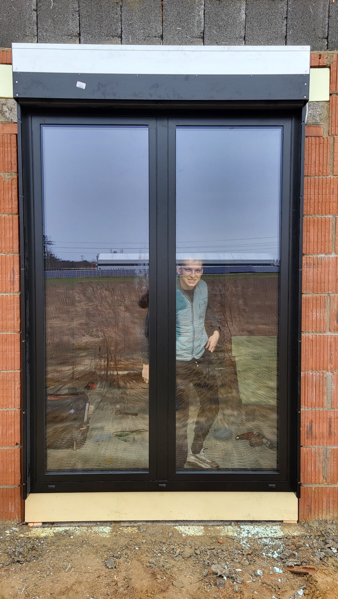 Czarne, dwuskrzydłowe okno z roletą zewnętrzną w surowym murze. W szybie odbija się uśmiechnięta osoba i narzędzia budowlane.