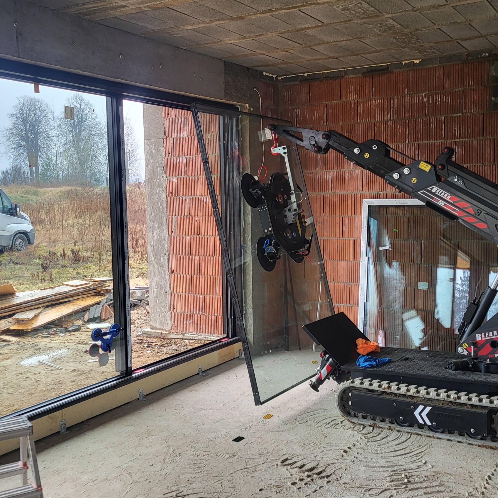 Montaż dużego okna z użyciem specjalistycznego robota na gąsienicach w surowym wnętrzu budynku. Widoczne przyssawki mocujące szybę.