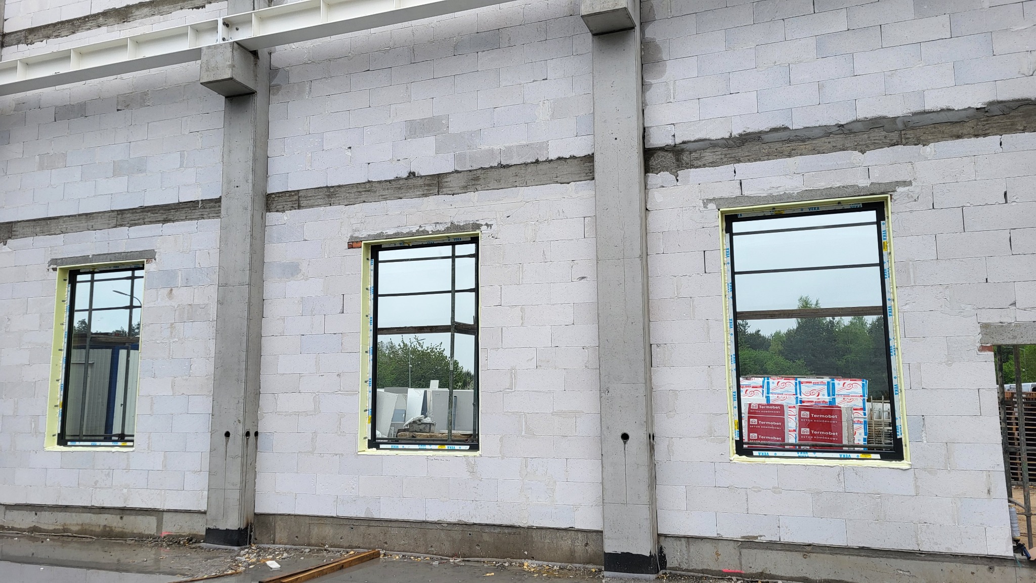 Montaż okien z czarną ramą w surowym murze z betonu komórkowego, widoczne paczki materiałów budowlanych. Żółta pianka montażowa wokół okien.