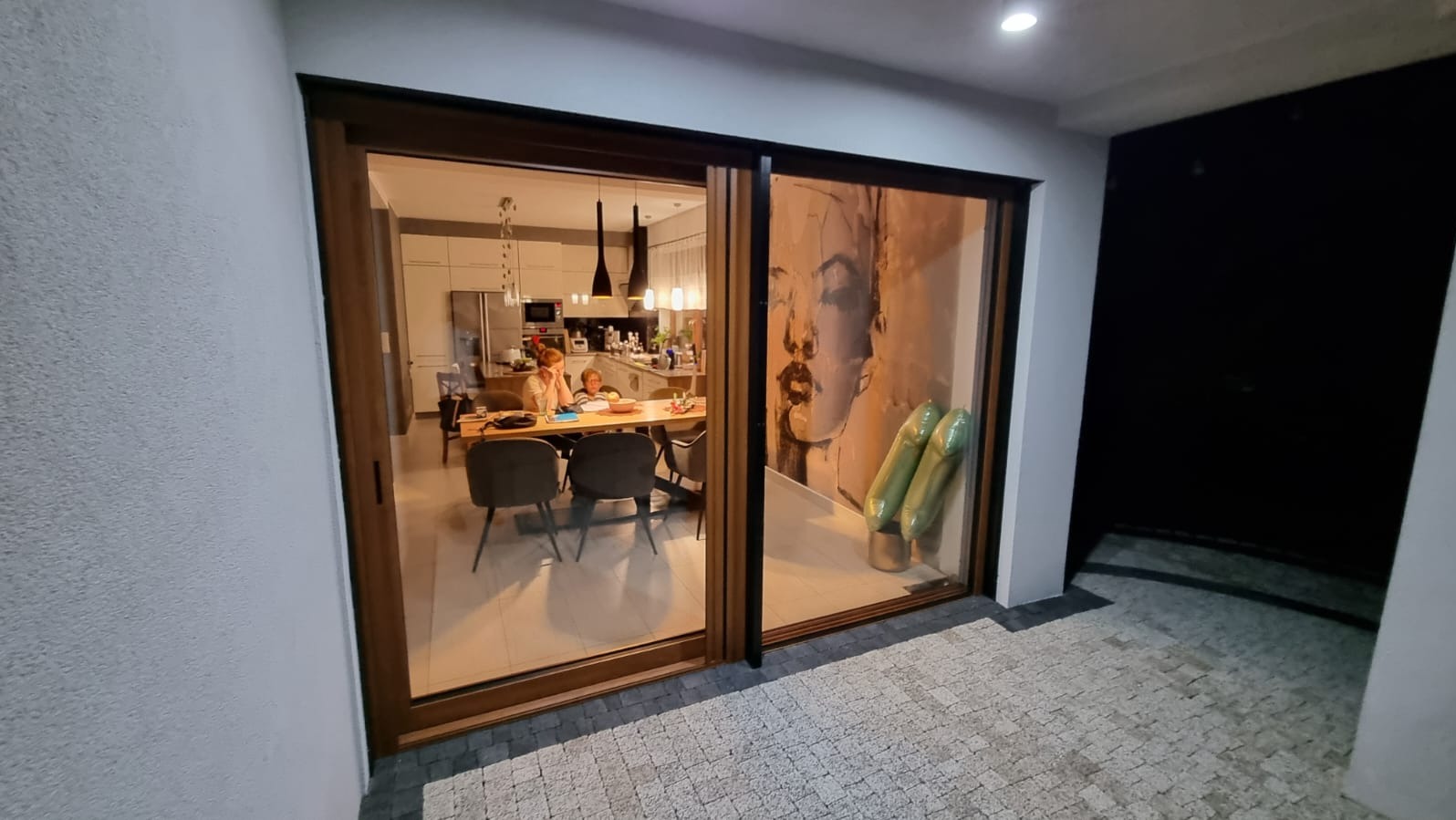 Drewniane drzwi przesuwne z widokiem na wnętrze domu: stół, krzesła, osoby. Po prawej stronie drzwi widoczna grafika. Na zewnątrz kostka brukowa.