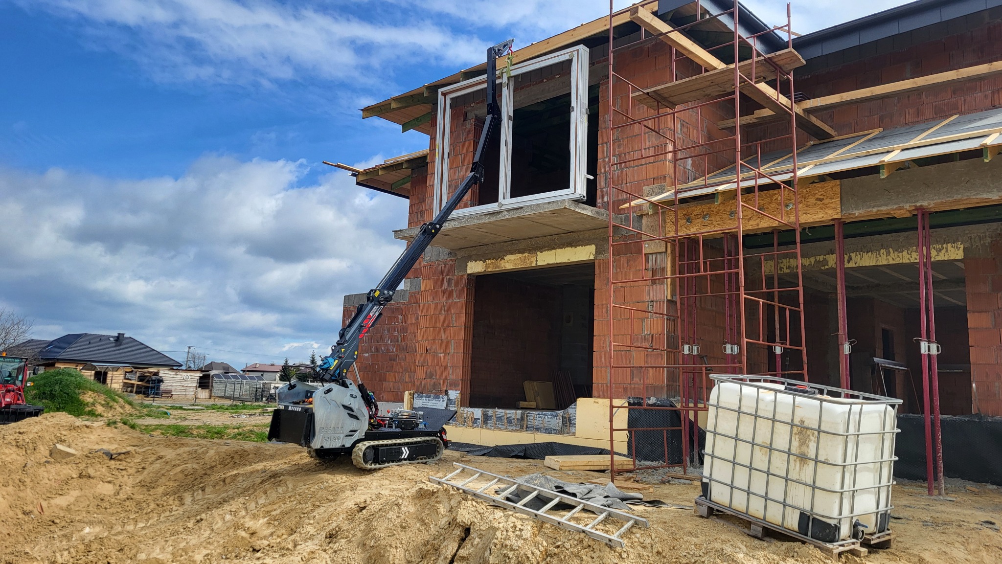 Montaż okna na budowie z użyciem mini żurawia na gąsienicach. Budynek z czerwonej cegły, rusztowania. Błękitne niebo z chmurami w tle.