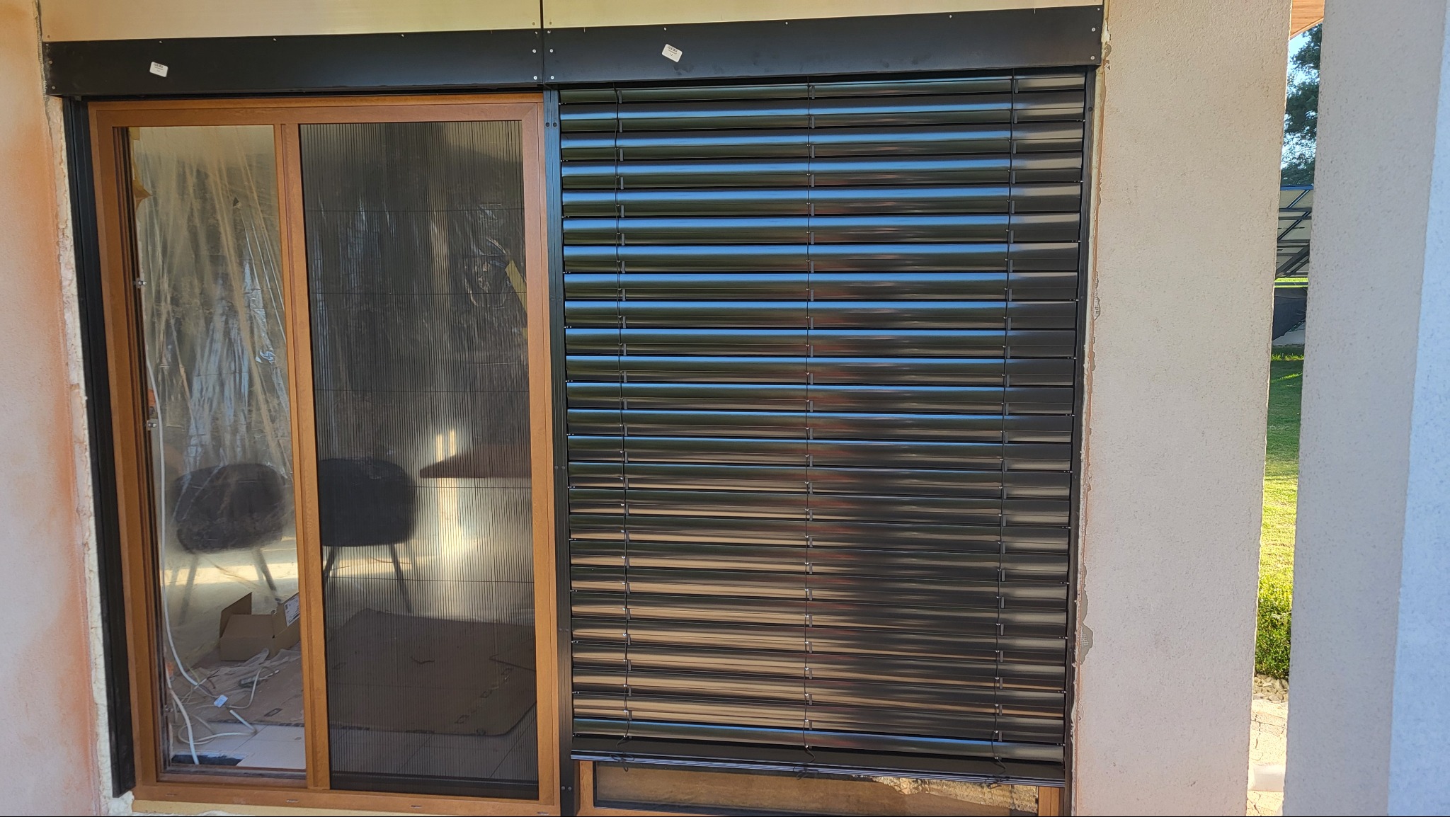 Ciemne rolety zewnętrzne zamontowane obok okna z moskitierą. Widoczne niedokończone prace wykończeniowe ściany wokół okna i rolet.