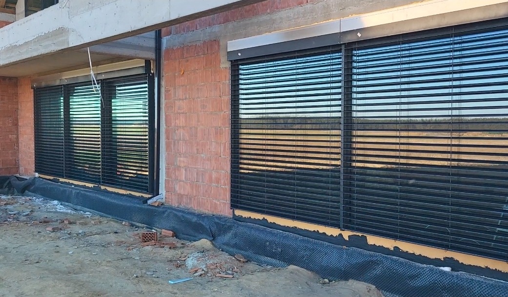 Ciemne rolety zewnętrzne zamontowane w oknach budynku w trakcie budowy. Widoczne cegły i surowy beton. Rolety z poziomymi lamelami odbijają krajobraz.