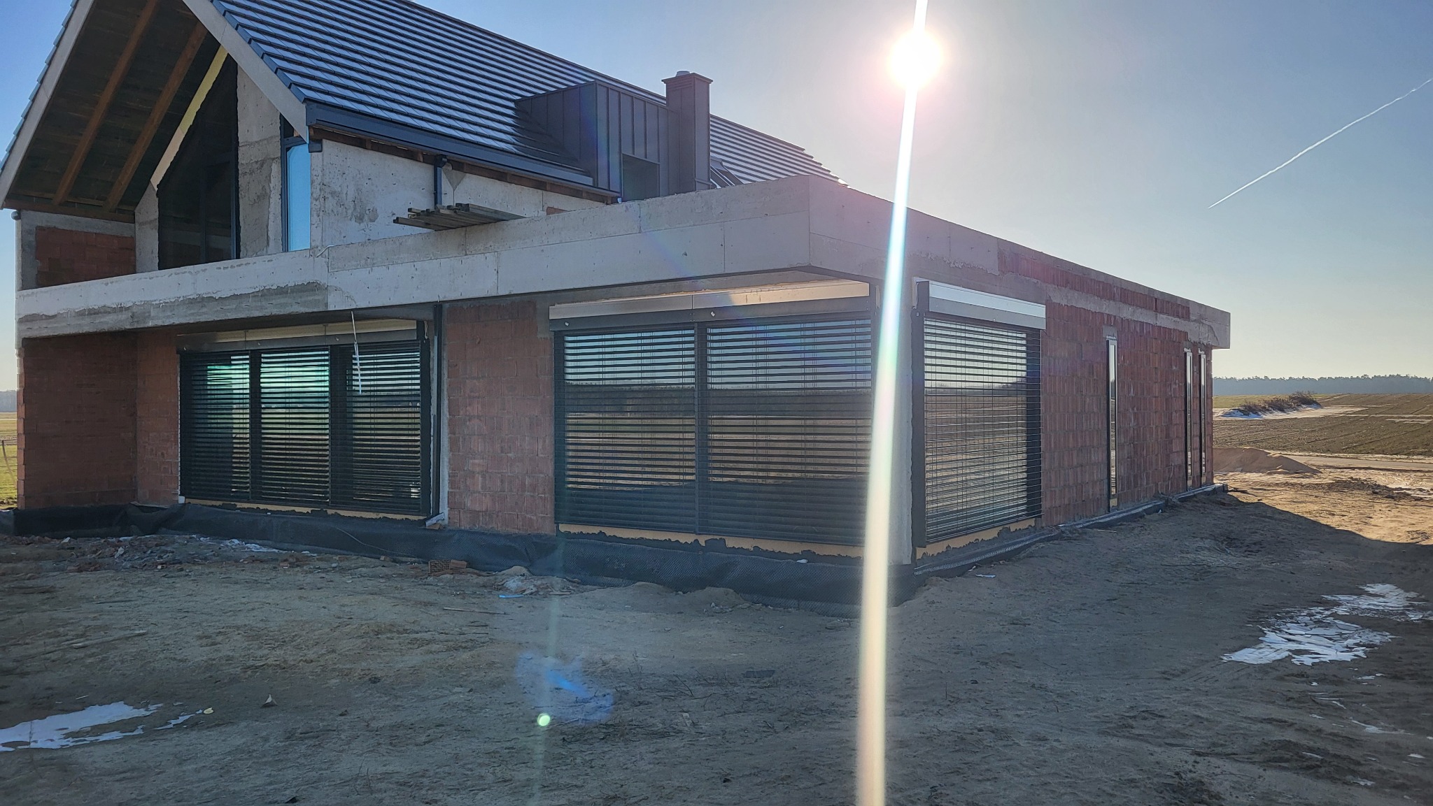 Surowe mury domu z zamontowanymi roletami zewnętrznymi w kolorze antracytowym. Widoczne niedokończone elewacje i otoczenie placu budowy w słoneczny dzień.