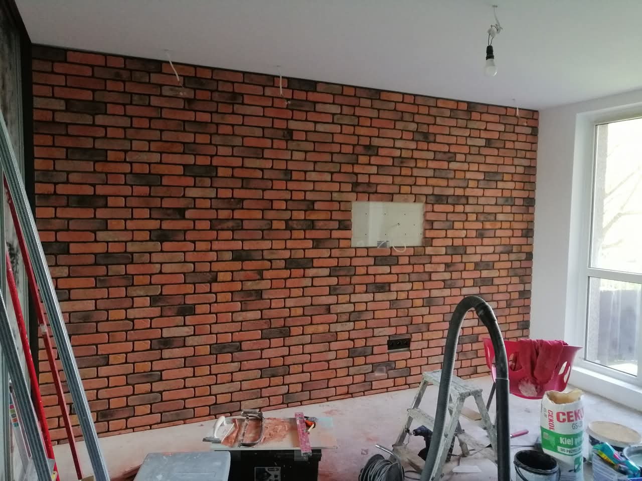 Ściana wyłożona dekoracyjną cegłą w odcieniach czerwieni i brązu, z widocznymi narzędziami i materiałami budowlanymi, sugerująca etap prac wykończeniowych we wnętrzu.