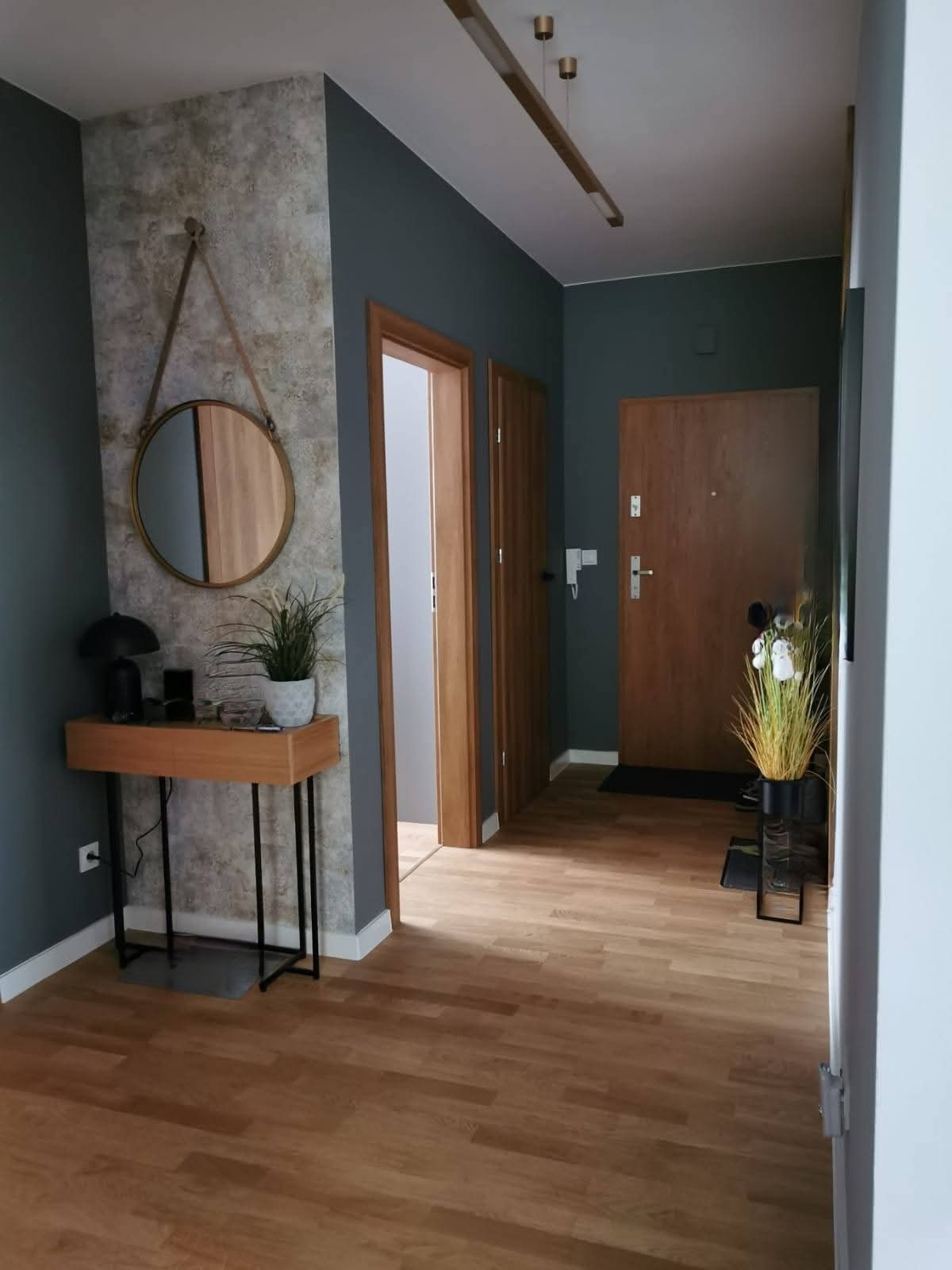 Nowoczesny przedpokój z drewnianą podłogą, szarą ścianą i okrągłym lustrem. Nad konsolą dekoracyjna tapeta imitująca kamień, minimalistyczne oświetlenie.