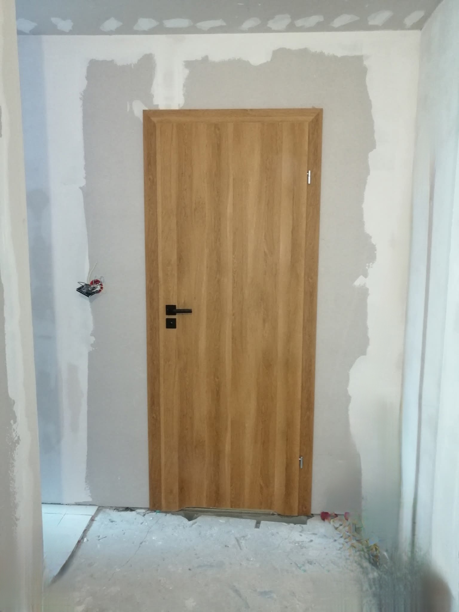 Montaż drzwi wewnętrznych z drewnianą okleiną. Ściany w trakcie szpachlowania, widoczna puszka elektryczna. Wykończenie wnętrza w trakcie realizacji.