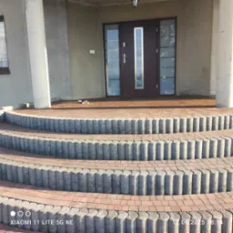 Schody wejściowe z kostki brukowej prowadzące do domu z brązowymi drzwiami. Trzy stopnie z obrzeżami z szarej kostki. Widok z dołu na elewację budynku.