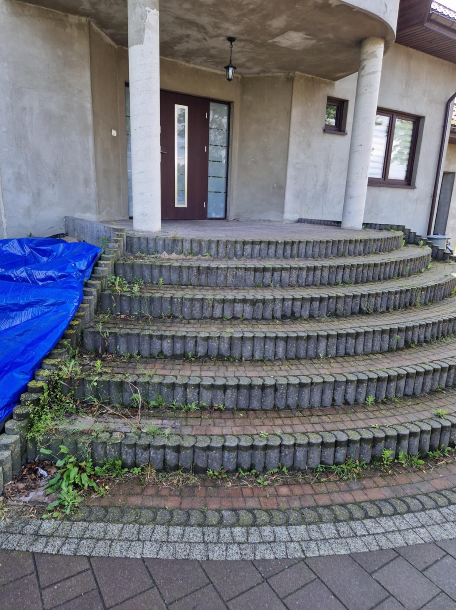 Schody z kostki brukowej przed wejściem do budynku z nieotynkowaną elewacją, częściowo zakryte niebieską plandeką, porośnięte zielenią.