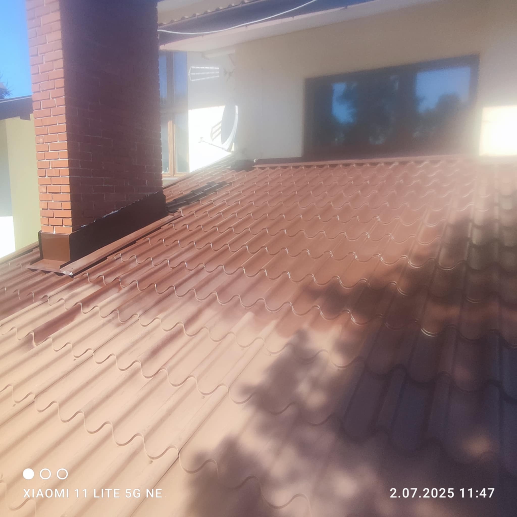 Błyszczący, brązowy dach z blachy falistej, widok z góry, komin z cegły w tle, okno w odbiciu. Nowe pokrycie dachowe.