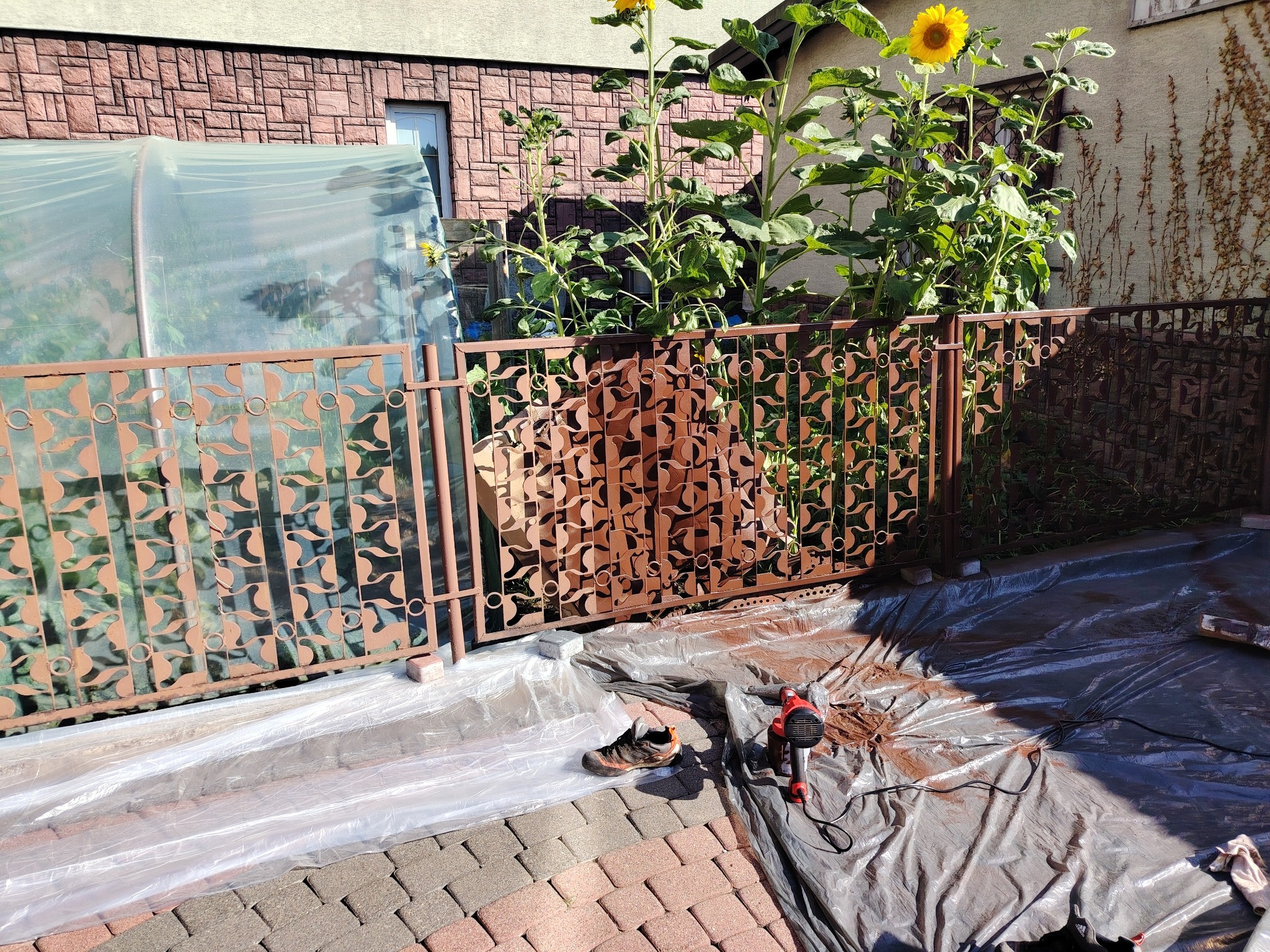 Malowanie metalowego ogrodzenia z dekoracyjnymi elementami. Widoczny pistolet natryskowy, folia ochronna i pojedynczy but roboczy. W tle roślinność i budynek.