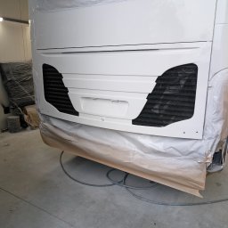 Piotr Lewandowski - Tył białego autokaru lub autobusu w warsztacie, świeżo polakierowany, z czarnymi kratkami wentylacyjnymi, częściowo zakryty folią ochronną i papierem przed dalszymi pracami.
