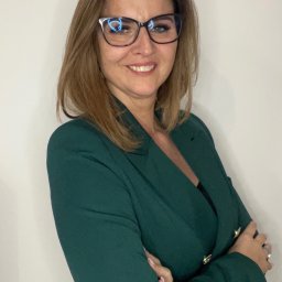 Żaneta Panek - Profesjonalny portret kobiety w okularach i zielonym żakiecie, z założonymi rękami na tle białej ściany. Uśmiech, pewny wyraz twarzy.