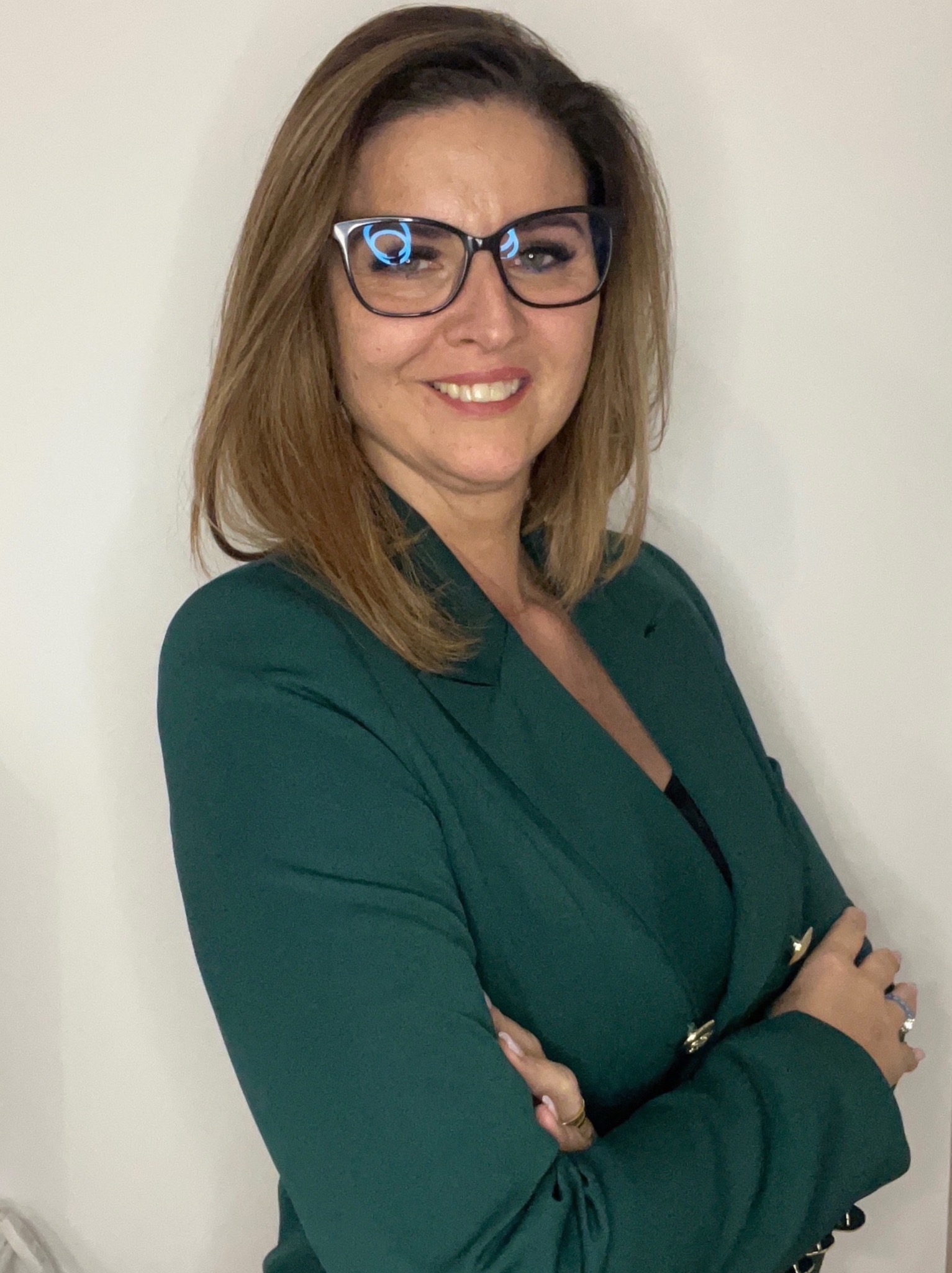 Profesjonalny portret kobiety w okularach i zielonym żakiecie, z założonymi rękami na tle białej ściany. Uśmiech, pewny wyraz twarzy.