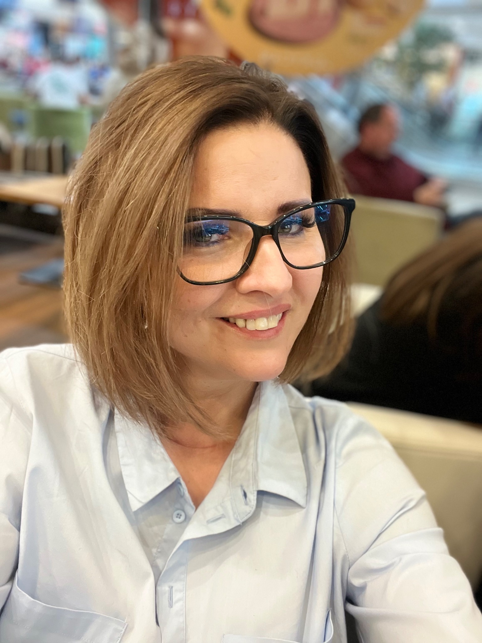 Uśmiechnięta kobieta w okularach, z krótkimi brązowymi włosami, w jasnej koszuli, siedząca w kawiarni. W tle widoczni inni ludzie.