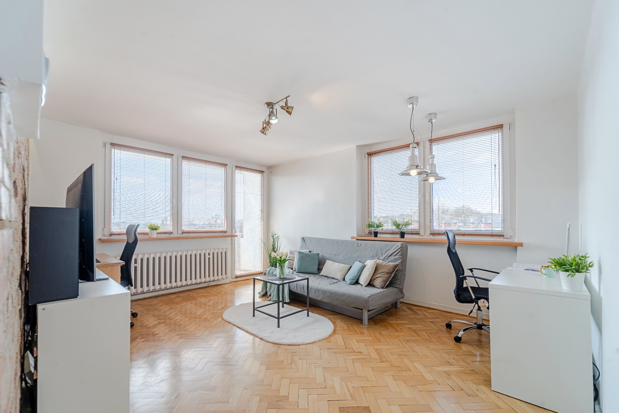 Jasne wnętrze mieszkania z parkietem w jodełkę, szarą sofą, biurkami i dużymi oknami z żaluzjami. Dwa stanowiska pracy z krzesłami biurowymi.