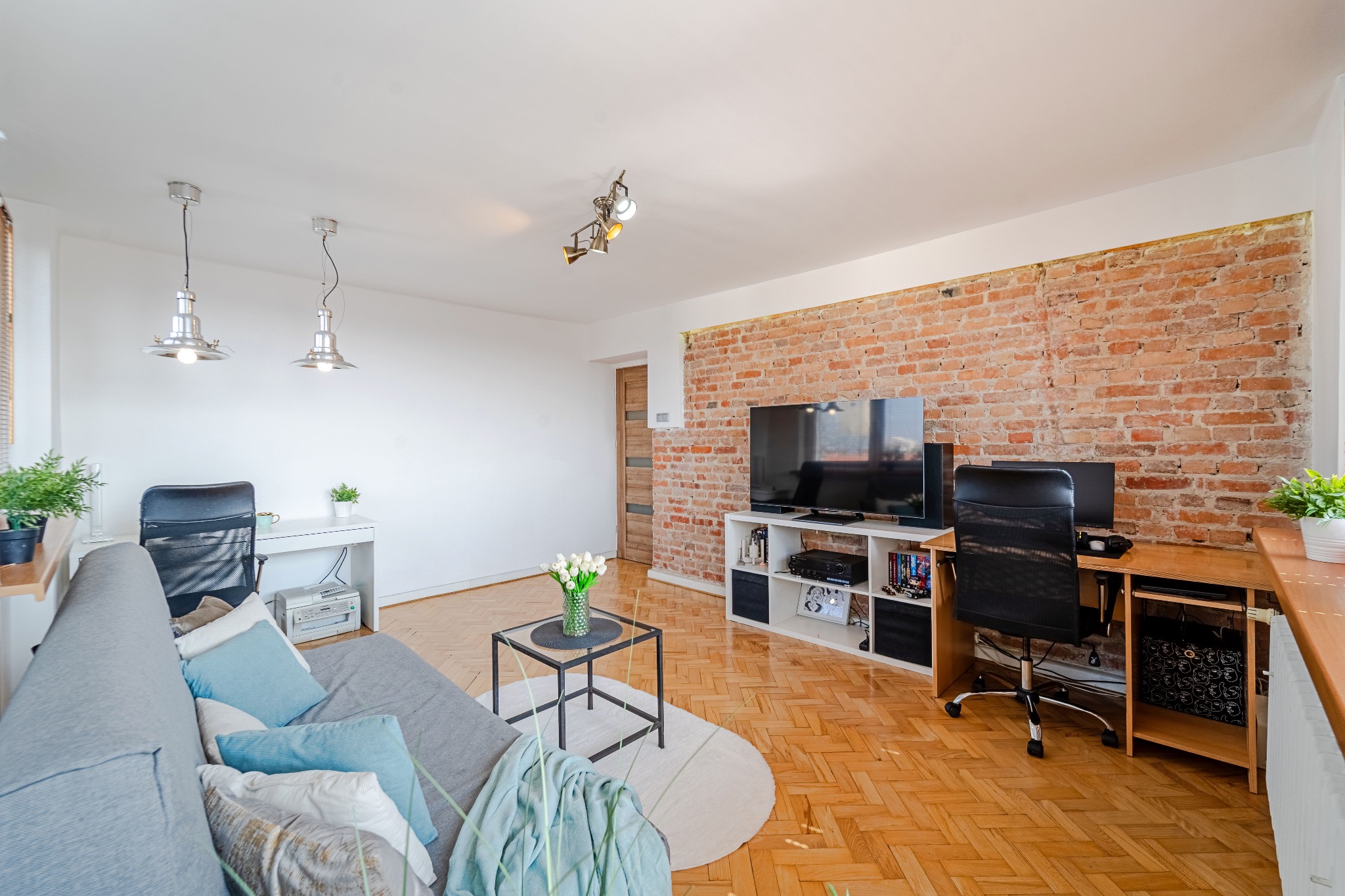 Jasne wnętrze mieszkania z ceglaną ścianą, drewnianą podłogą i nowoczesnym wyposażeniem: biurko, sofa z poduszkami, telewizor i stolik z kwiatami.