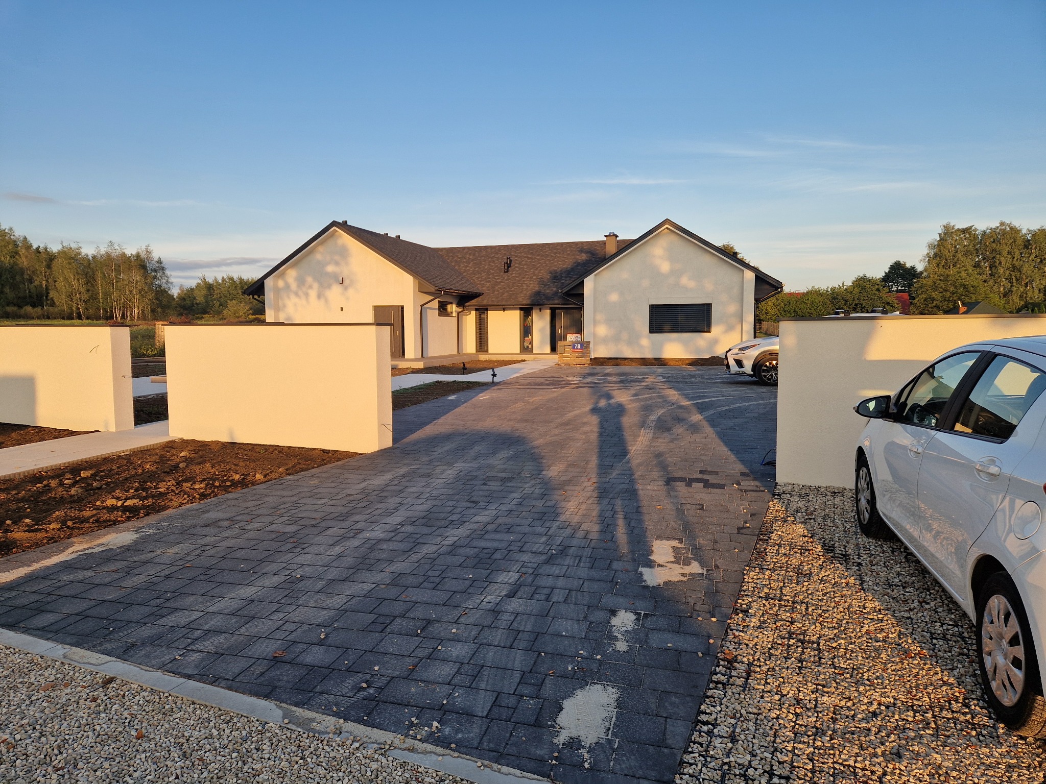 Nowoczesny podjazd z szarej kostki brukowej prowadzący do domu jednorodzinnego. Widoczne samochody i cień osoby fotografującej. Całość oświetlona ciepłym światłem zachodzącego słońca.