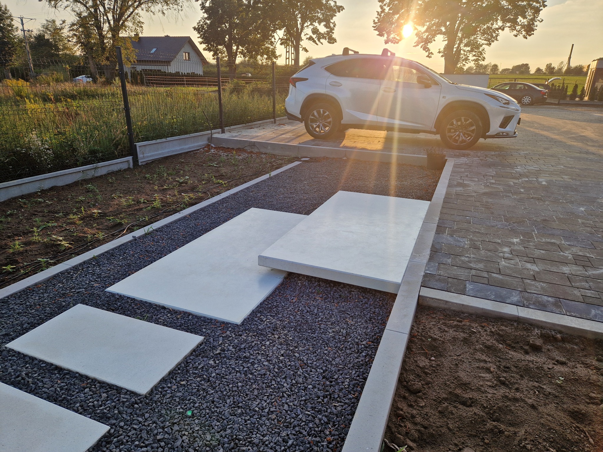 Nowoczesny podjazd z szarego żwiru i betonowych płyt w ogrodzie, z białym SUV-em w tle, oświetlony promieniami zachodzącego słońca.