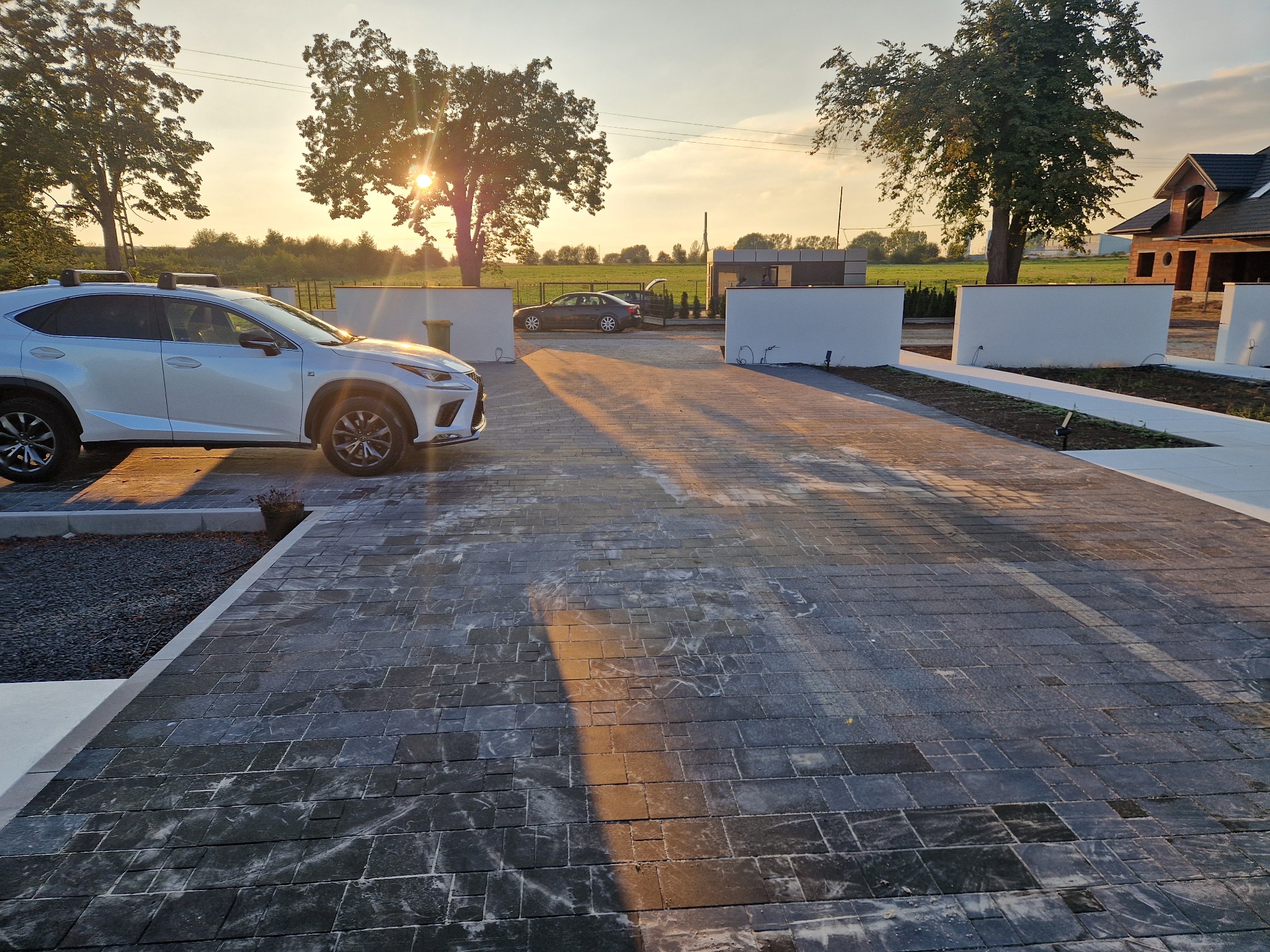 Nowoczesny podjazd z ciemnej kostki, oświetlony zachodzącym słońcem. Widoczny samochód Lexus i fragment domu w budowie w tle. Minimalistyczne ogrodzenie.