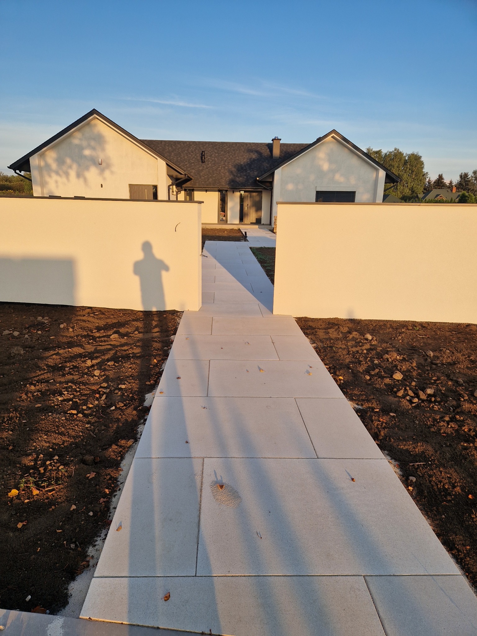 Nowoczesna ścieżka z jasnych płyt prowadząca do domu, otoczona białymi murkami. Cień osoby na murze. Minimalistyczny design, świeżo ułożona nawierzchnia.