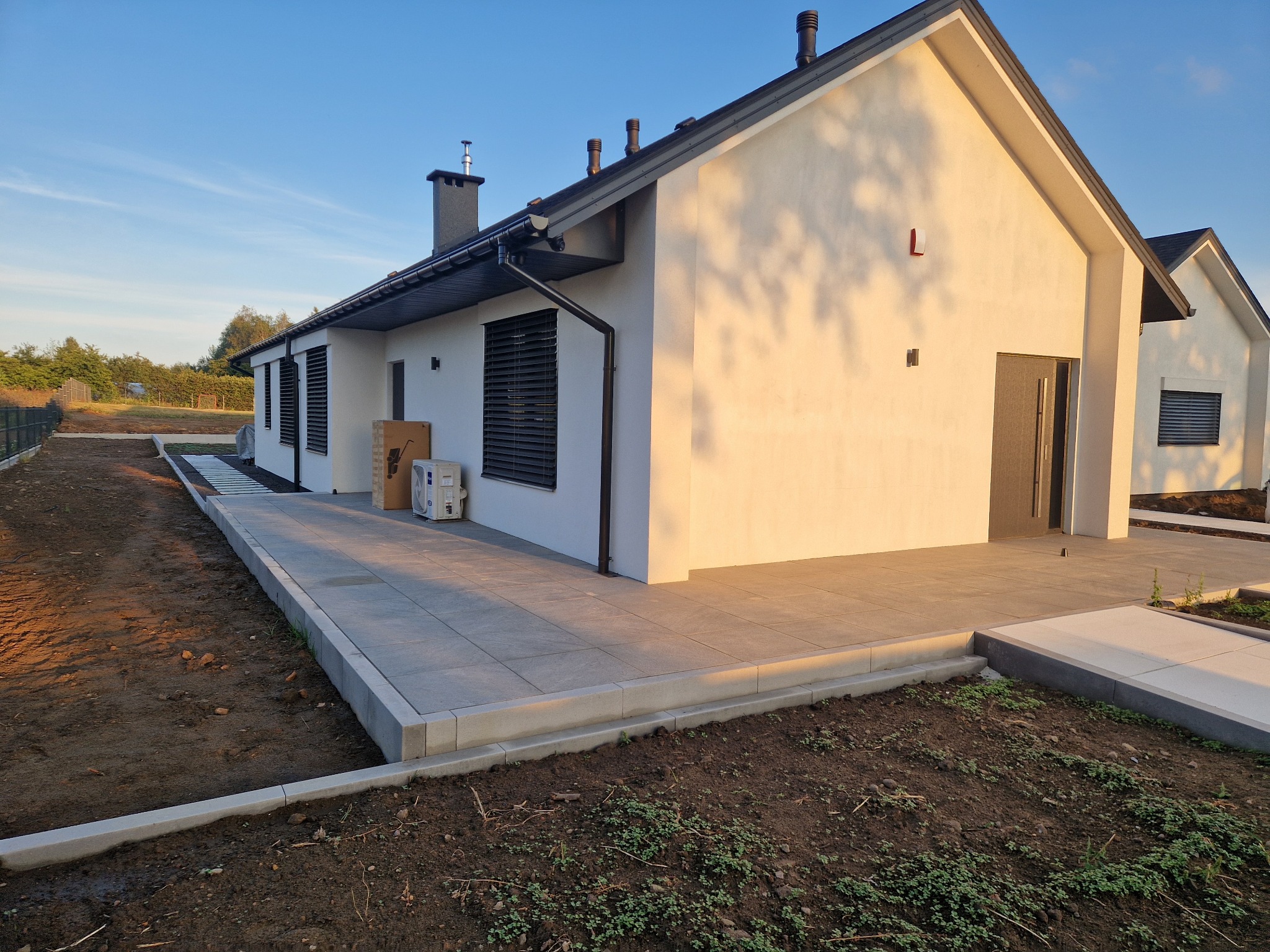 Nowoczesny dom jednorodzinny z jasną elewacją i tarasem wyłożonym szarą kostką. Widoczne obrzeża i przygotowany teren pod ogród. Błękitne niebo w tle.