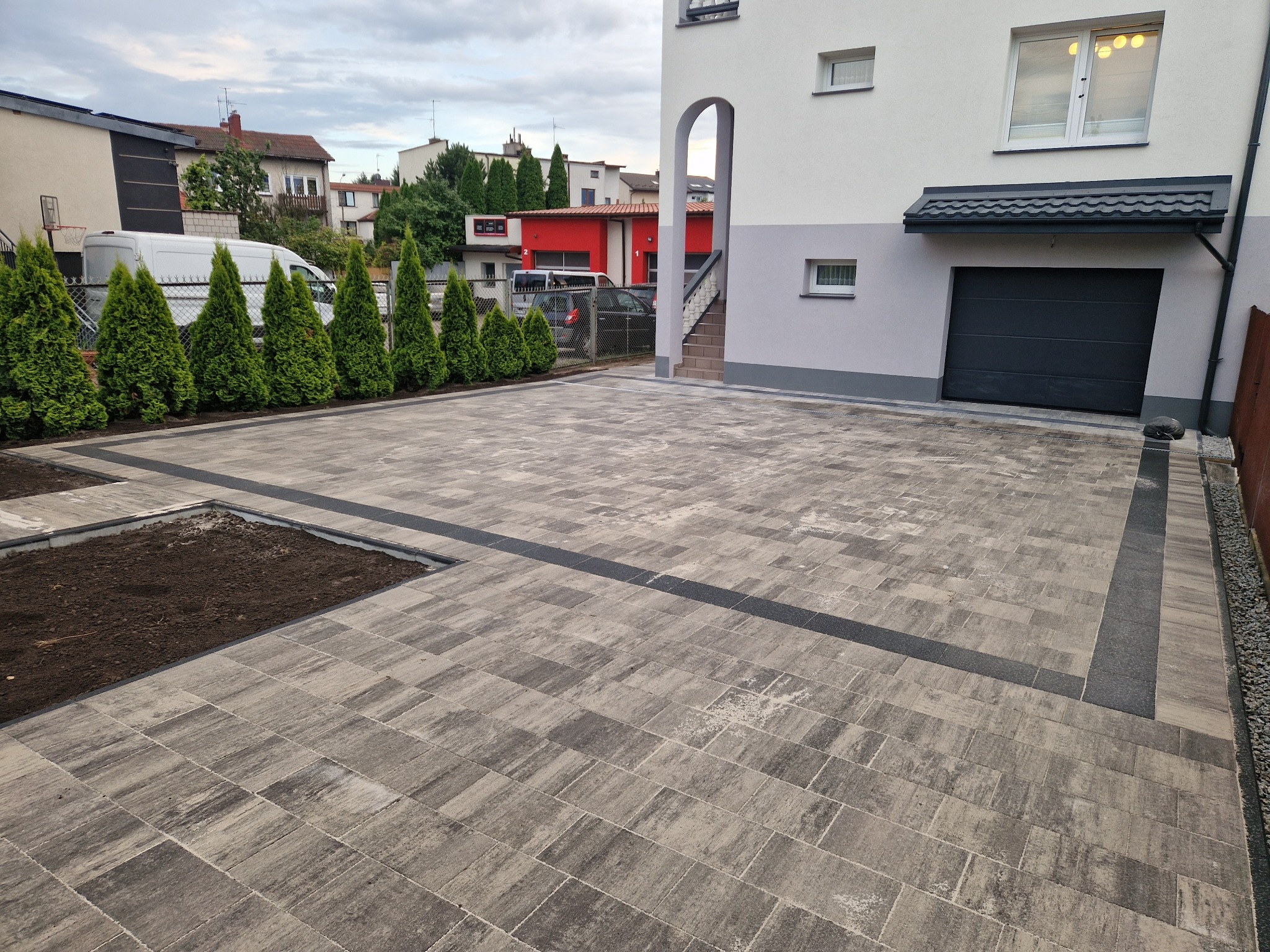 Nowoczesny podjazd z szarej kostki brukowej ułożonej we wzór jodełki, obramowany ciemniejszą kostką. Widoczne tuje i budynek w tle.