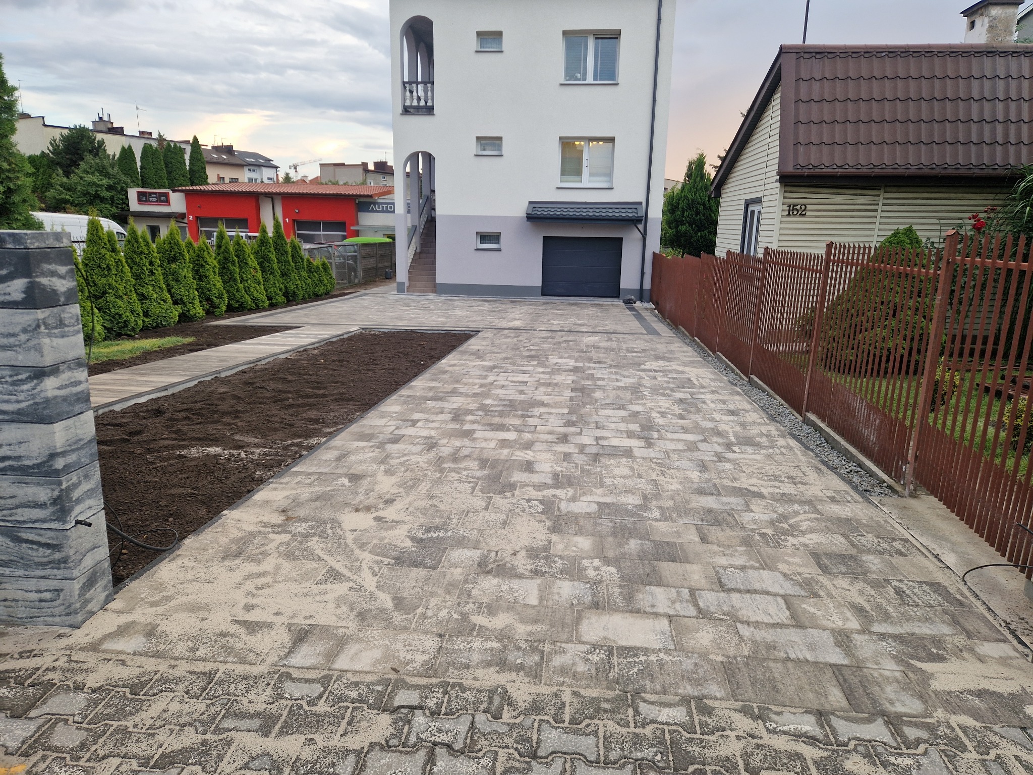 Nowa, szara kostka brukowa na podjeździe do domu z garażem. Widoczne rabaty ziemi po bokach i ogrodzenie. Ujęcie szerokokątne, podkreślające przestrzeń.