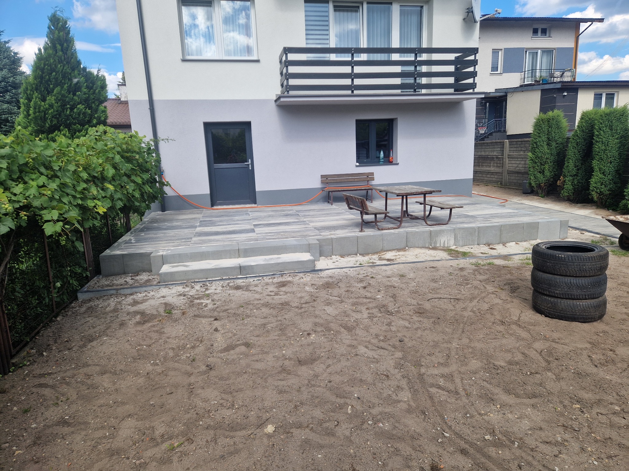 Nowoczesny taras z szarej kostki brukowej przy domu z balkonem. Widoczne schody z kostki, meble ogrodowe i przygotowany teren pod dalsze prace.