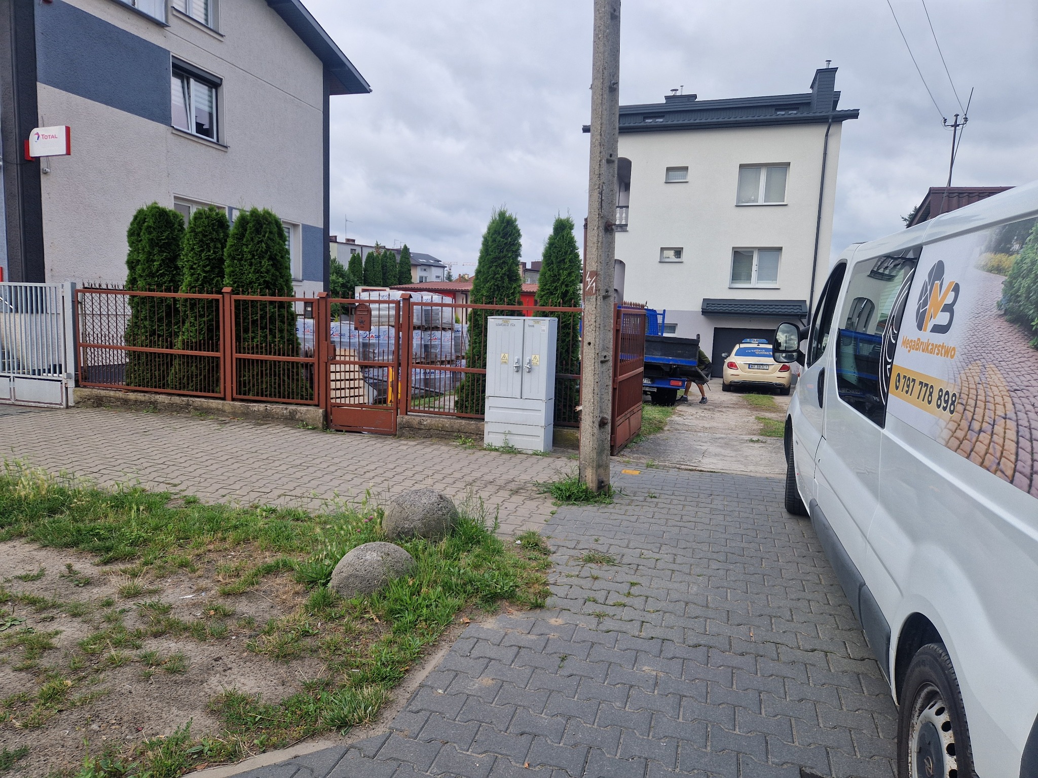 Ułożona szara kostka brukowa przed budynkiem z ogrodzeniem. Na pierwszym planie dwa kamienie i zaparkowany samochód dostawczy z reklamą firmy brukarskiej.