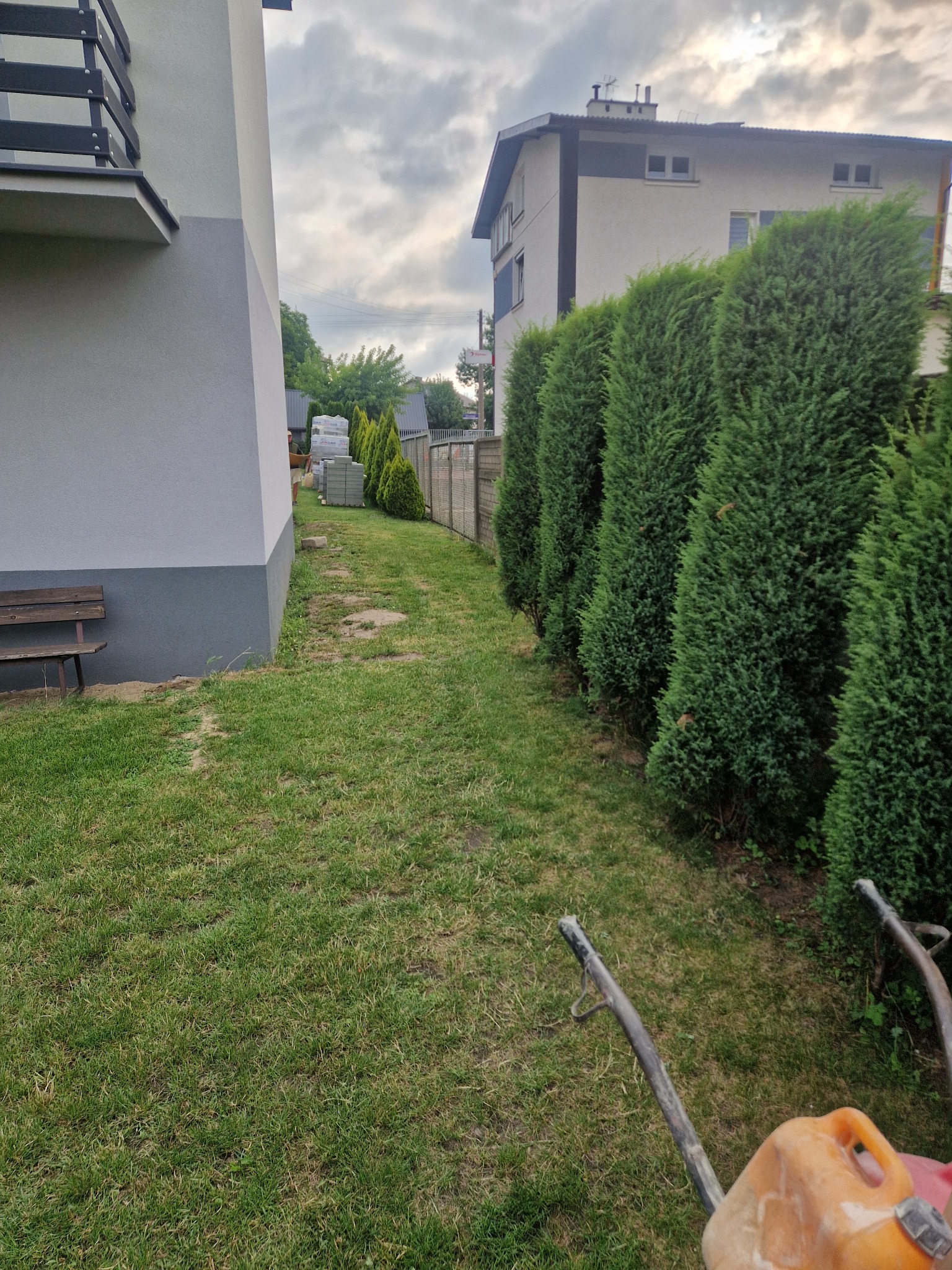 Przygotowany teren pod budowę ścieżki z kostki, widoczne materiały budowlane i narzędzia. Tuje wzdłuż ogrodzenia. Dom w tle.