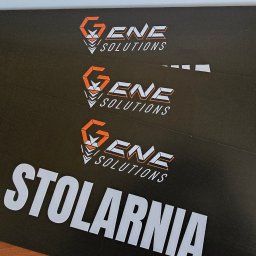 VisualLook Łukasz Rajkowski - Druk UV na czarnych płytach z logo firmy z Uniejowa i napisem 'Stolarnia', odporny na warunki atmosferyczne, widoczne na stole z bliska.