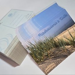 VisualLook Łukasz Rajkowski - Stos kartek pocztowych z nadrukiem plaży i napisem 'Pozdrowienia z Ustki', obok stos pocztówek z motywem żeglarskim, logo 'Fregata'.