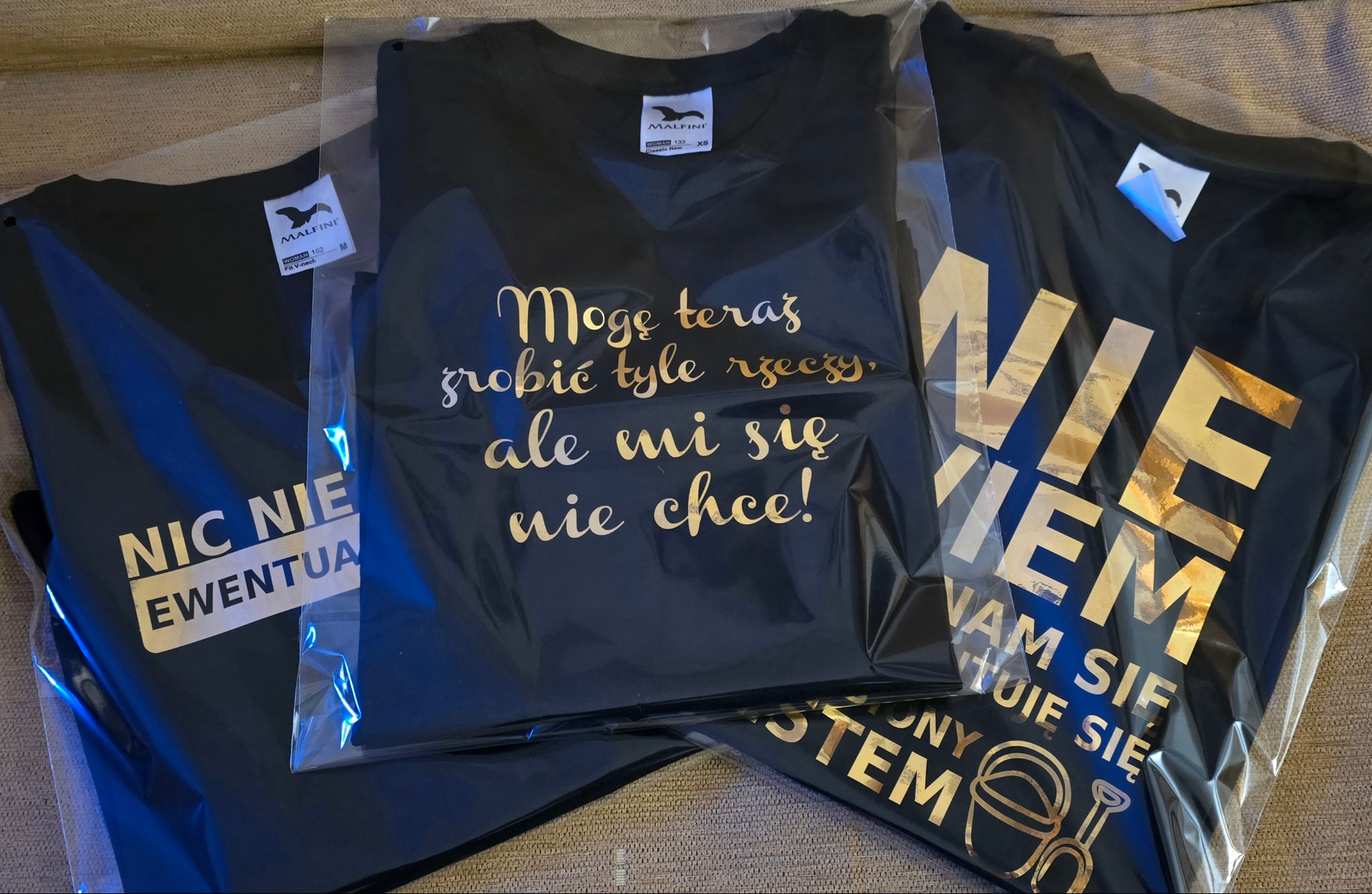 Trzy czarne koszulki w folii z nadrukami w kolorze złotym: „Nic nie ewentualnie”, „Mogę teraz robić tyle rzeczy, ale mi się nie chce!”, „Nie wiem, nam się tuje się”.