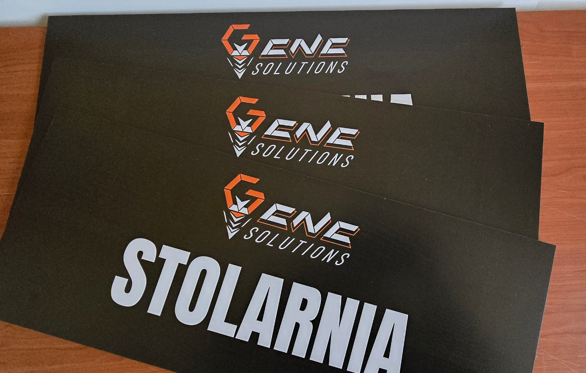 Druk UV na czarnych płytach z logo firmy z Uniejowa i napisem 'Stolarnia', odporny na warunki atmosferyczne, widoczne na stole z bliska.