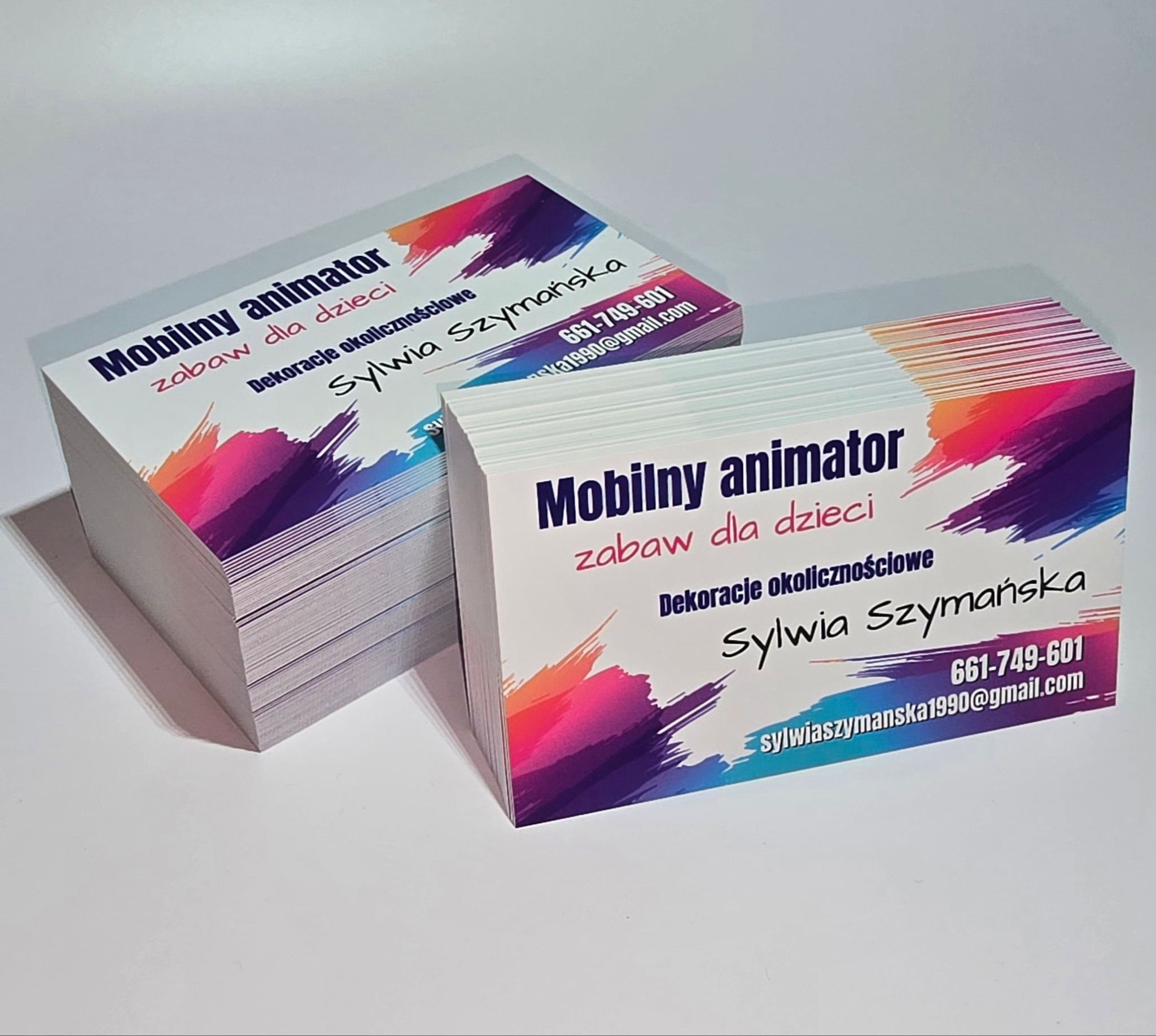 Dwa stosy wizytówek z kolorowym motywem, na których widnieje napis: Mobilny animator, zabawy dla dzieci, dekoracje okolicznościowe, Sylwia Szymańska, numer telefonu i adres email.