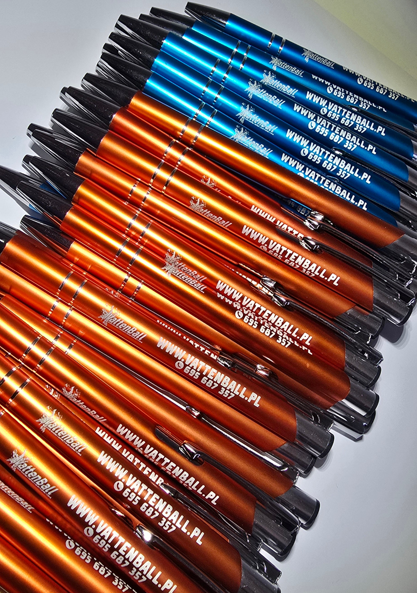 Rząd niebieskich i pomarańczowych długopisów z nadrukiem logo i danych kontaktowych firmy, ułożonych ukośnie na jasnym tle. Gadżety reklamowe.
